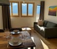 Bright living and dining area with wooden floors, large windows offering mountain views, a grey sofa, a dining table set for two, and a painting on the wall.