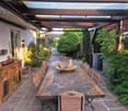 An outdoor patio with a wooden table and chairs, surrounded by lush greenery and a covered wooden structure.