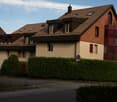 Two-story house with a brown tiled roof, multiple windows, and a red balcony section. The house is surrounded by greenery and a hedge, with a parking area in front.