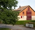 Two-story house with a red-tiled roof, beige walls, and red shutters. Features include a balcony on the upper floor and a well-maintained hedge in the front yard.