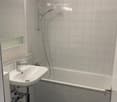 Modern bathroom with a white sink, bathtub, and glass shelf. Features grey tiles on walls and black tiles on the floor.
