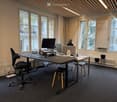 Office space with a large desk, multiple computer screens, ergonomic chairs, and a small side table with a cup. Large windows with a view of a balcony and outdoor area.