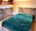 Cozy attic bedroom with wooden beams, a bed covered in a teal duvet, a small stool, and a speaker on a nightstand. The walls feature a large, leafy mural.