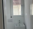 Modern bathroom with a white wall-mounted sink, chrome faucet, and a toilet. A window with a view of a tiled roof is above the sink.