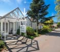 A modern residential building entrance with a covered porch, mailboxes, and a bicycle parked nearby. The area is surrounded by greenery and trees, with a clear pathway leading to the entrance.