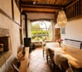 Cozy dining area with a large wooden table, chairs with fur cushions, a stone fireplace, and a chandelier hanging from the wooden ceiling.