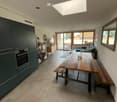 Open-plan kitchen and dining area with a large wooden dining table, benches, a sofa, dark green cabinets, and a modern oven. Large windows and a skylight provide natural light.