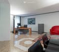 Modern office space with a red sofa, black leather chairs, a wooden desk with a laptop and screen, a glass coffee table, and a large painting on the wall.