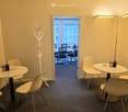 Modern, minimalist room with white walls, carpeted floor, and a coat rack. Two round tables with chairs are arranged, and a floor lamp is positioned near the doorway leading to another room.