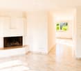 Bright hallway with tiled flooring, white walls, and a built-in fireplace on one side. A window at the end allows natural light to enter, revealing greenery outside.