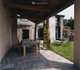 Outdoor patio area with a wooden dining table and chairs, surrounded by a stone building and greenery.