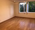Empty room with wooden flooring and large window with open shutters, showing greenery outside.