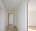 Modern, minimalist hallway with white walls, light wooden flooring, and recessed ceiling lighting. Features a closed door and an open doorway leading to another room.