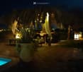 Nighttime outdoor area with a swimming pool, stone-paved ground, large thatched-roof structure, tables and chairs, decorative lanterns, and potted plants.