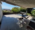 A modern, tiled balcony with a table and chairs, overlooking a garden and distant sea view.