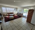 Bright living room with tiled floor, large windows, and sliding glass doors leading to a balcony. Features a brown sofa, coffee table, ottoman, and a small wooden chair.