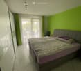 Bright bedroom with green walls, a large bed with patterned bedding, a nightstand with a lamp, and a wardrobe with mirrored doors. Natural light filters through sheer curtains covering a window that leads to a balcony.