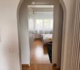 A narrow hallway with wooden flooring and light-colored walls, leading to a bedroom with a bed, rug, and window with curtains.
