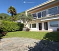 A well-maintained garden with a stone pathway leading to a modern house. The garden features lush green grass, a wooden bench, and a lawn mower. There are various plants and trees, including a palm tree, and the house has large windows and a balcony.