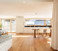 Open-plan living and dining area with light wood flooring, white walls, and large windows overlooking a snowy mountain view. Features a sofa, dining table with chairs, kitchen area with cabinets and a sink, and a potted plant near the window.
