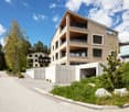 Modern multi-story building with large windows and balconies, surrounded by greenery and trees.