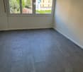 Empty room with grey tiled flooring, white walls, and large windows with blinds overlooking a green area.
