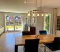 Bright, open-plan living and dining area with wooden flooring, large windows, a sofa, a dining table with chairs, and a cabinet with decorative items.