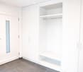Empty room with white built-in wardrobes, shelves, and a hanging rod, featuring a grey tiled floor and a white door with a frosted glass panel.