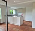 Modern kitchen with white cabinets, a central island, and terracotta tiled flooring. Features a sink with a faucet and large windows allowing natural light.