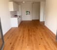 Empty room with light wooden flooring and white walls, featuring a small kitchen area with cabinets and a sink.