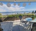 Balcony with a scenic view of a lake and mountains, featuring a round glass table, chairs, a wicker chair, and potted plants.