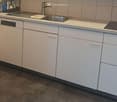 Modern kitchen with white cabinets, stainless steel sink, and dishwasher. Grey tiled backsplash and dark grey concrete-style flooring.