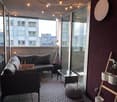 Modern balcony with dark grey sofa, coffee table, round stool, patterned rug, and potted plants. Large glass sliding doors open to a city view.