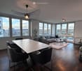 Modern living and dining area with large windows offering a city view, featuring a grey sectional sofa, a coffee table, a dining table with chairs, and a small bar cart with bottles.