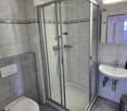 Modern bathroom with a glass-enclosed shower, toilet, sink, and mirror. The walls are tiled with a decorative border, and the floor is tiled with gray tiles.