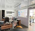Modern office space with wooden desk, ergonomic chair, computer setup, filing cabinet, sink, and framed artwork. Large windows provide natural light.
