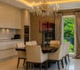 Elegant dining area with a dark wooden dining table surrounded by cream-colored chairs, modern cabinetry, and a chandelier. A wine cooler is visible near the window, and the room features patterned wallpaper and large windows with curtains.