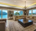 Elegant living room with large windows offering a scenic mountain and lake view, featuring a beige sectional sofa, dark wooden coffee table, armchair, chandelier, and decorative mirror.