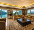 Elegant living room with large windows offering a scenic mountain and lake view, featuring a beige sectional sofa, dark wooden coffee table, armchair, chandelier, and decorative mirrors.