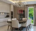 Modern kitchen and dining area with white cabinets, a dark dining table surrounded by white chairs, a chandelier, and a view of greenery through large windows.