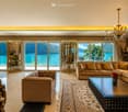 Elegant living room with large windows offering a scenic lake view, featuring a beige sectional sofa, coffee table, chandelier, and decorative mirrors.