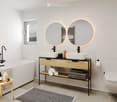 Modern bathroom with a freestanding bathtub, double sink vanity with two round mirrors, a small wooden stool, and a toilet. The space features white tiles, a grey rug, and a window with a view.