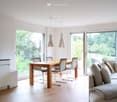 Bright, open-plan living and dining area with large windows, a modern sofa, a dining table with chairs, and pendant lamps.