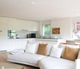 Modern open-plan living and dining area with a white sofa, glass coffee table, kitchen area with white cabinets and appliances, and a dining table with chairs.