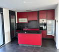 Modern kitchen with red cabinets, black countertops, and stainless steel appliances including a refrigerator and oven. Features a central island with a black countertop and a sink.