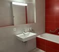 Modern bathroom with a white sink, chrome faucet, large mirror with built-in lighting, bathtub with red tiles, and a toilet. Features a handrail for accessibility.