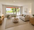 Modern living room with light-colored furniture, large windows, and a view of greenery outside. Features a sofa, armchair, coffee table, side tables, lamps, and decorative vases.