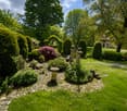 Well-maintained garden with neatly trimmed hedges, flowering shrubs, a stone pathway, and a central stone bench. The garden features a variety of plants and trees, with a lawn and a small fountain in the background.