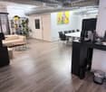 Modern office space with a sofa, coffee table, chairs, a large table, printer, and a small kitchen area with a coffee machine and cups.