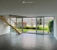 Open-plan space with a wooden staircase leading to an upper level, large glass doors opening to a garden, and a tiled floor.